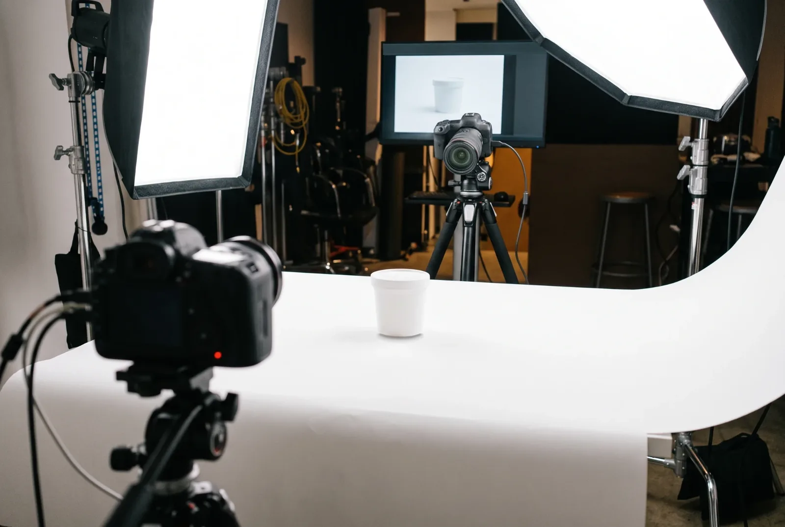 Fotostudio mit weißem Hohlkehlen-Hintergrund, einem weißen Becher in der Mitte, zwei Softboxen, zwei Kameras auf Stativen und einem Monitor im Hintergrund, der den Becher zeigt