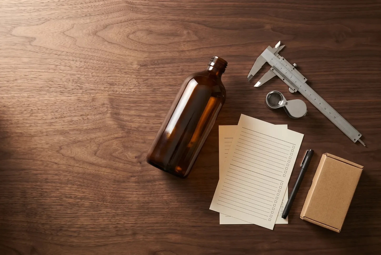 Walnussholz-Tisch von oben mit einer Braunglasflasche, Messschieber, Lupe, Prüfbögen mit Kästchen, Stift und kleinem Karton