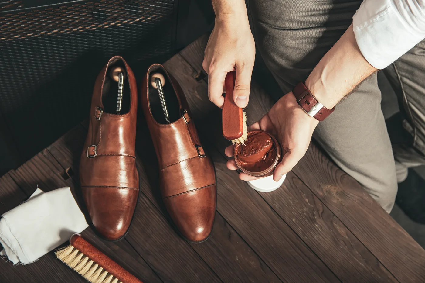 Hände pflegen ein Paar braune Lederschuhe mit Bürste und Creme auf einem Holzboden